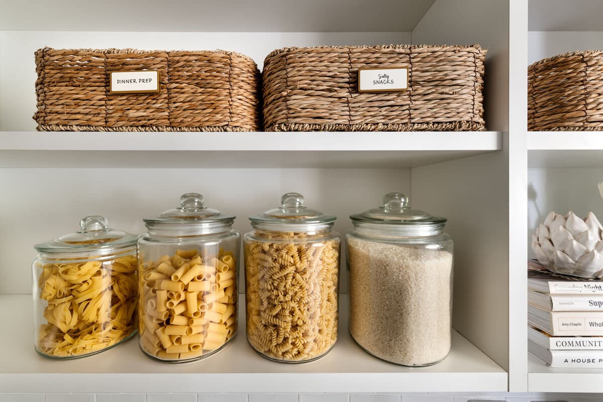 Labelled woven baskets on a pantry shelf above glass jars holding dried pasta and rice.