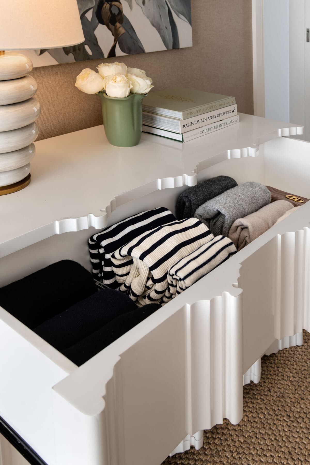 A shelf with stacked ceramics, white flowers in a green vase and books, above folded knitwear and a black and cream striped throw.