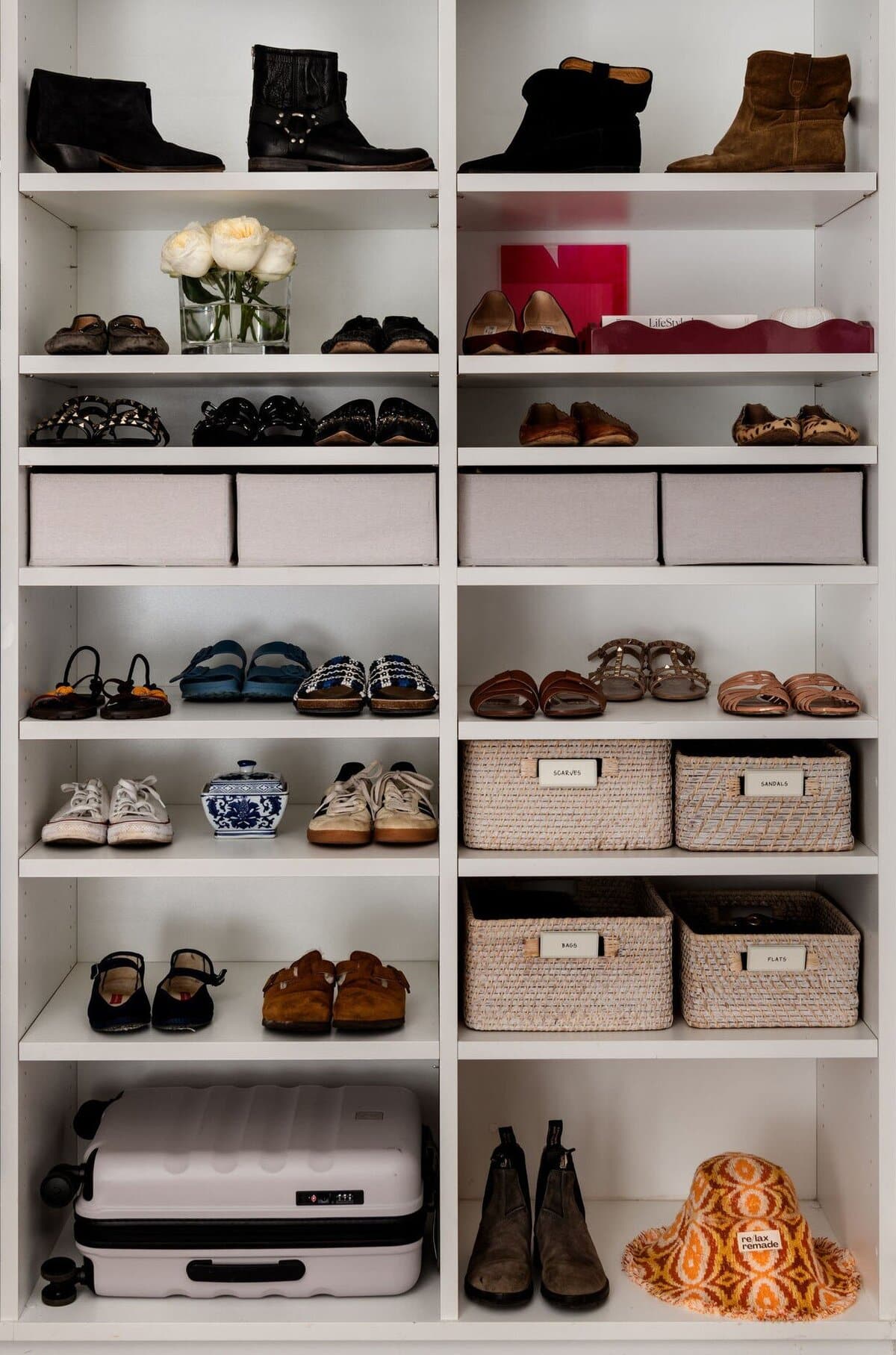 Floor to ceiling white shelving holding shoes arranged in pairs, with labelled fabric boxes and woven baskets on the upper shelves.